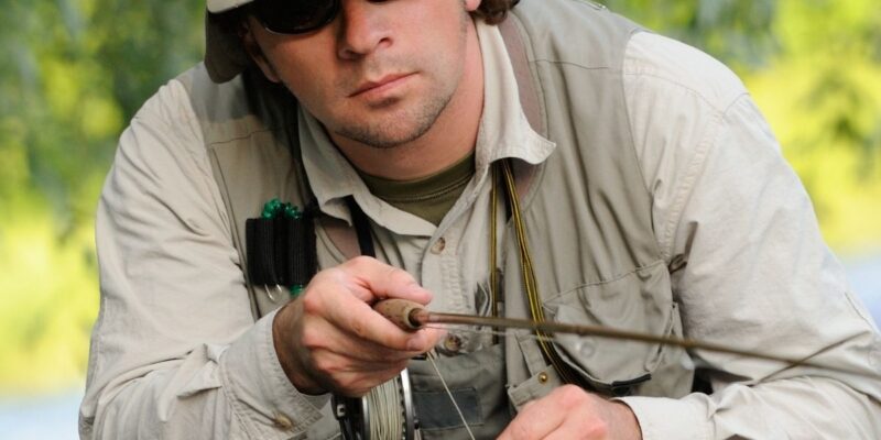 a fly fisherman wearing a khaki fly fishing hat with neck flap holding a fly fishing pole and pulling in fishing line