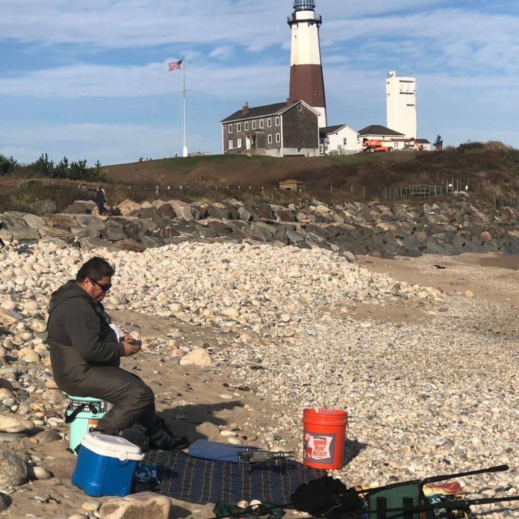 Surf fisherman in waders preparing to fish from the rocky Montauk shoreline with the Montauk Lighthouse in the background.